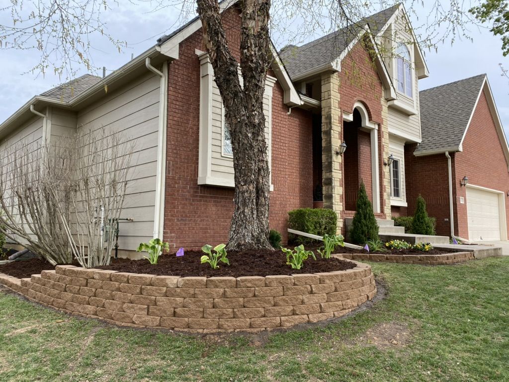 New stone retaining wall bed with fresh mulch and hostas wrapped around a tree at the front of the home
