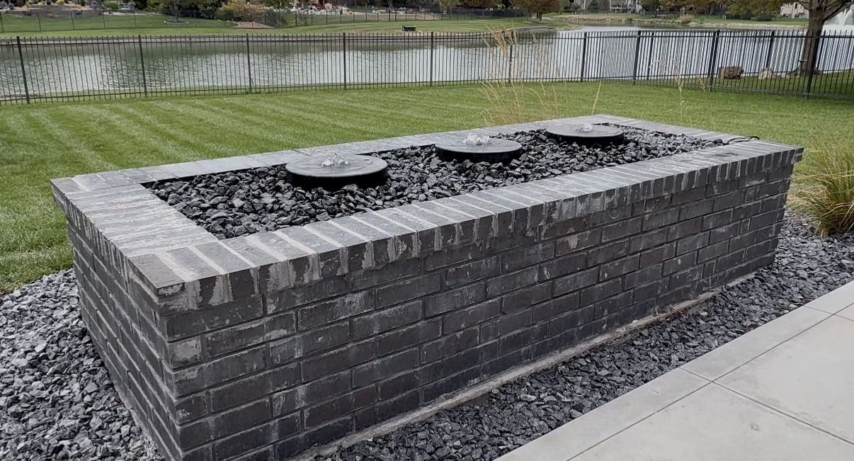 Brick basin filled with black rock and three round granite bubblers, with a lake in the background